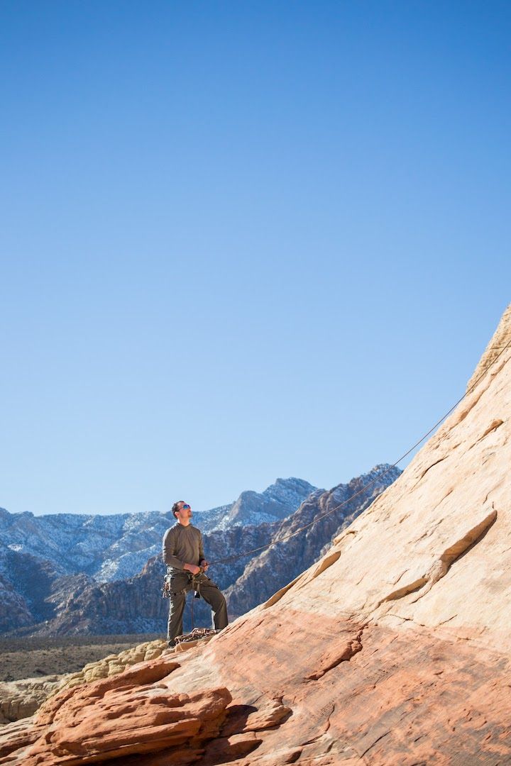 Man belaying/holding a rope on red rocks, with snow covered mountains in the background below a clear blue sky.